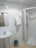 Un baño con un lavamanos blanco, un espejo, una ducha con puerta de vidrio y un cubo de basura azul.