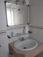 Un baño con lavamanos blanco, espejo y repisa de vidrio con artículos de aseo.