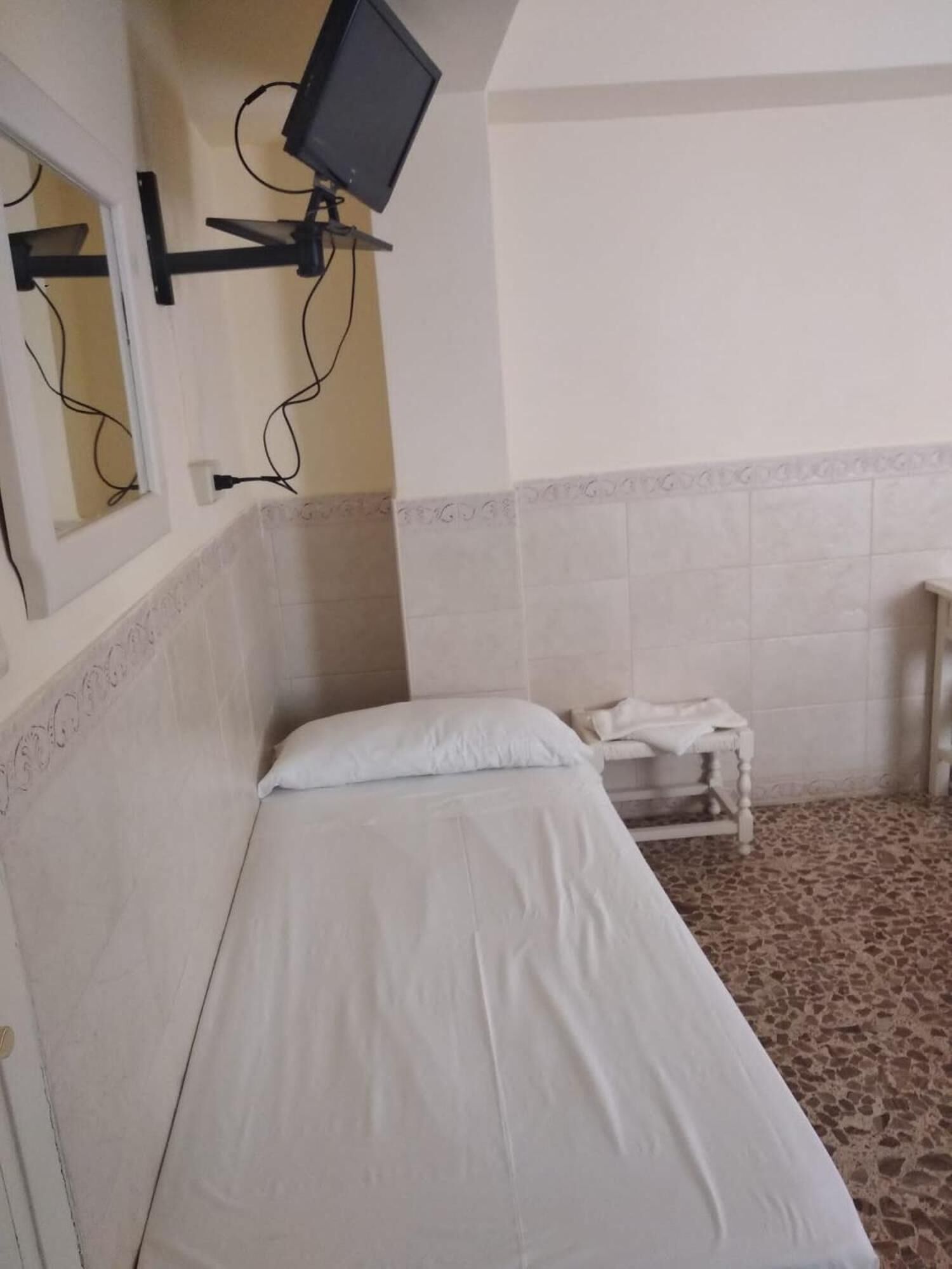 Una habitación pequeña con cama individual, televisión montada en la pared, una mesita y una silla.
