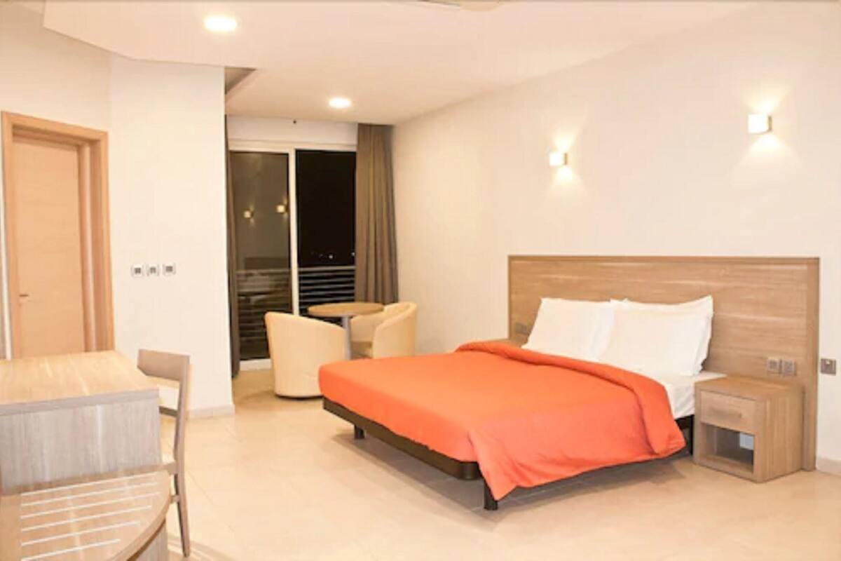 A modern hotel room with a large bed, a small table, a chair, and a view of the city at night.
