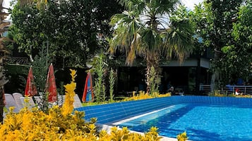 Una piscina rodeada de abundante vegetación y palmeras, con sombrillas rojas y sillas de descanso blancas.