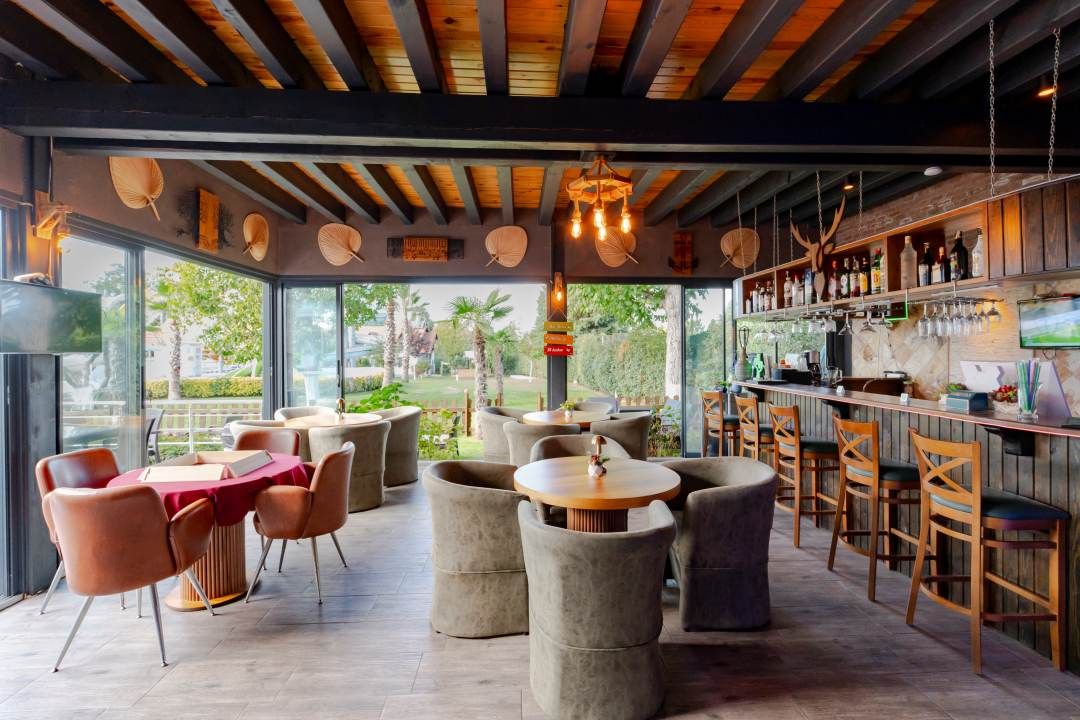 A modern bar area with round tables and chairs, a wooden ceiling, and a view of greenery outside.