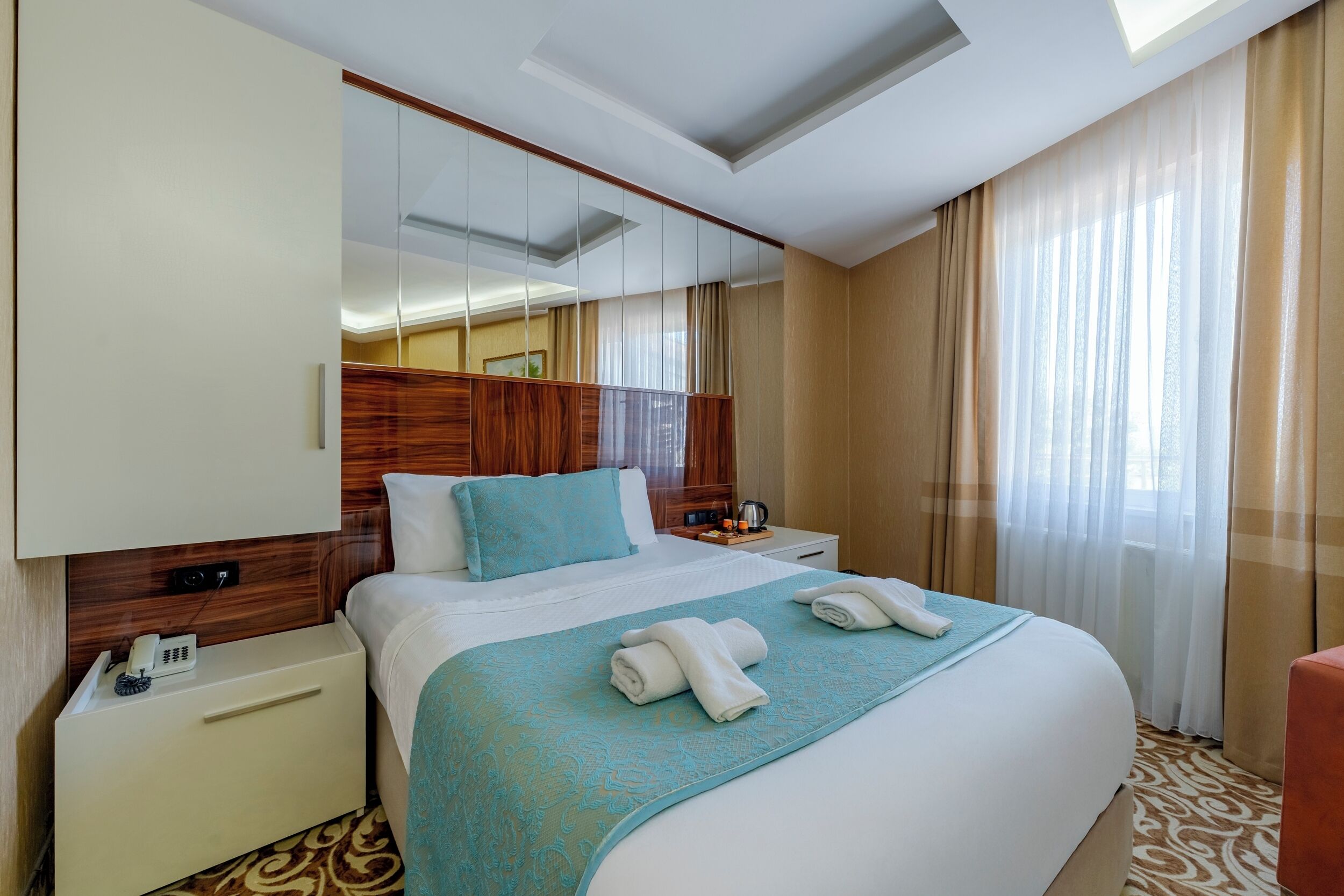 A hotel room with a large bed, a bedside table with a phone, a mirror, and a window with curtains.