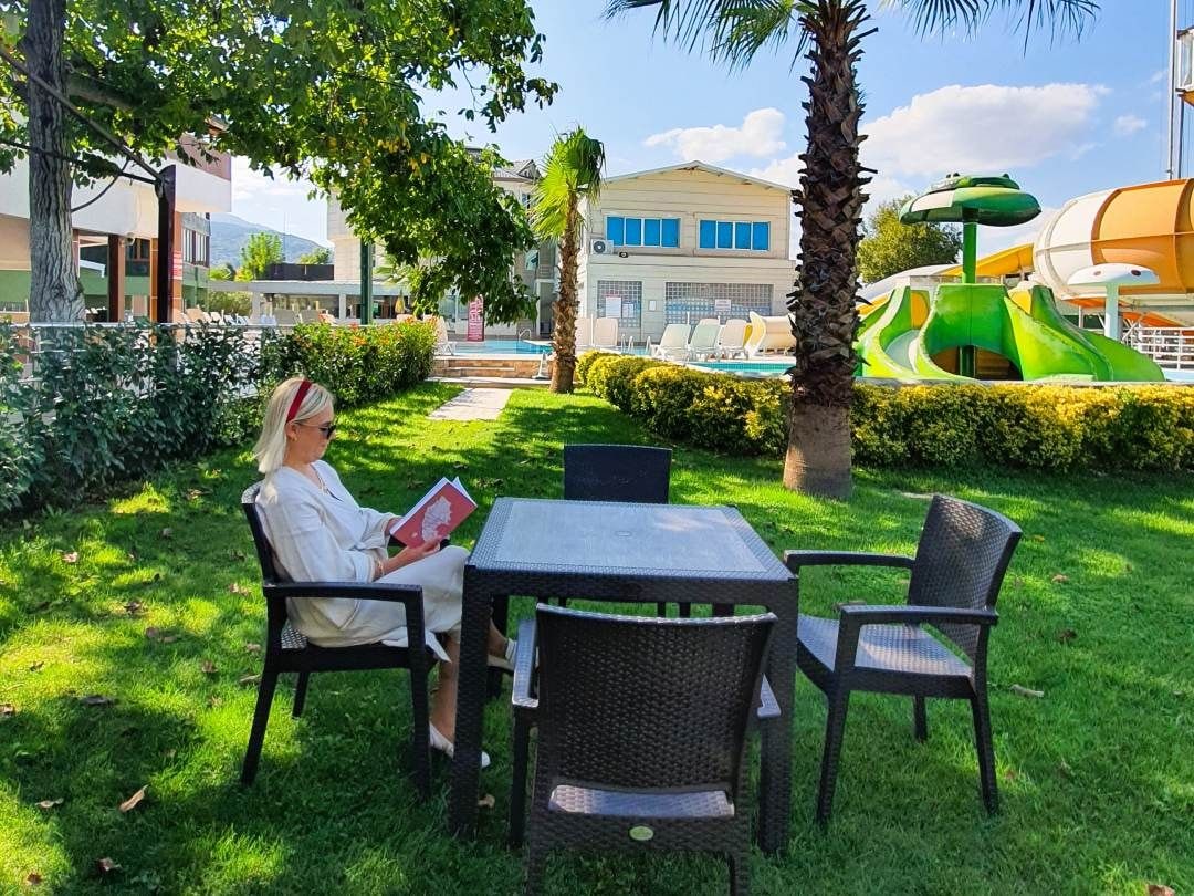 A person sitting at a table in a grassy outdoor area with a pool and water slides in the background.
