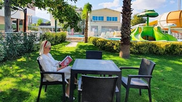 A person sitting at a table in a grassy outdoor area with a pool and water slides in the background.