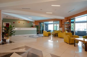Una hall d'albergo con reception, area salotto con sedie gialle e una grande vetrata che permette l'ingresso di luce naturale.