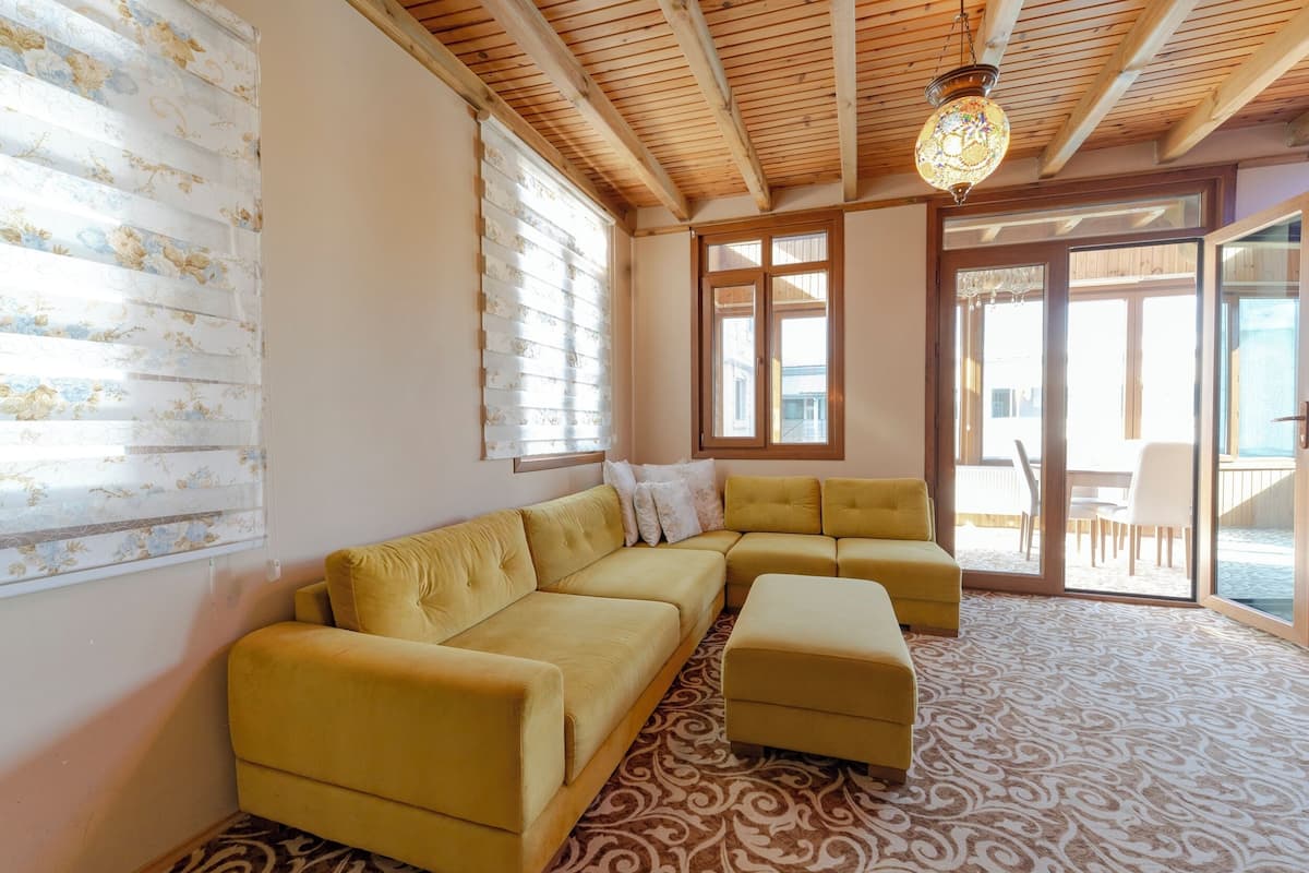 A living room with a yellow sectional sofa, patterned carpet, wooden ceiling beams, and large windows.