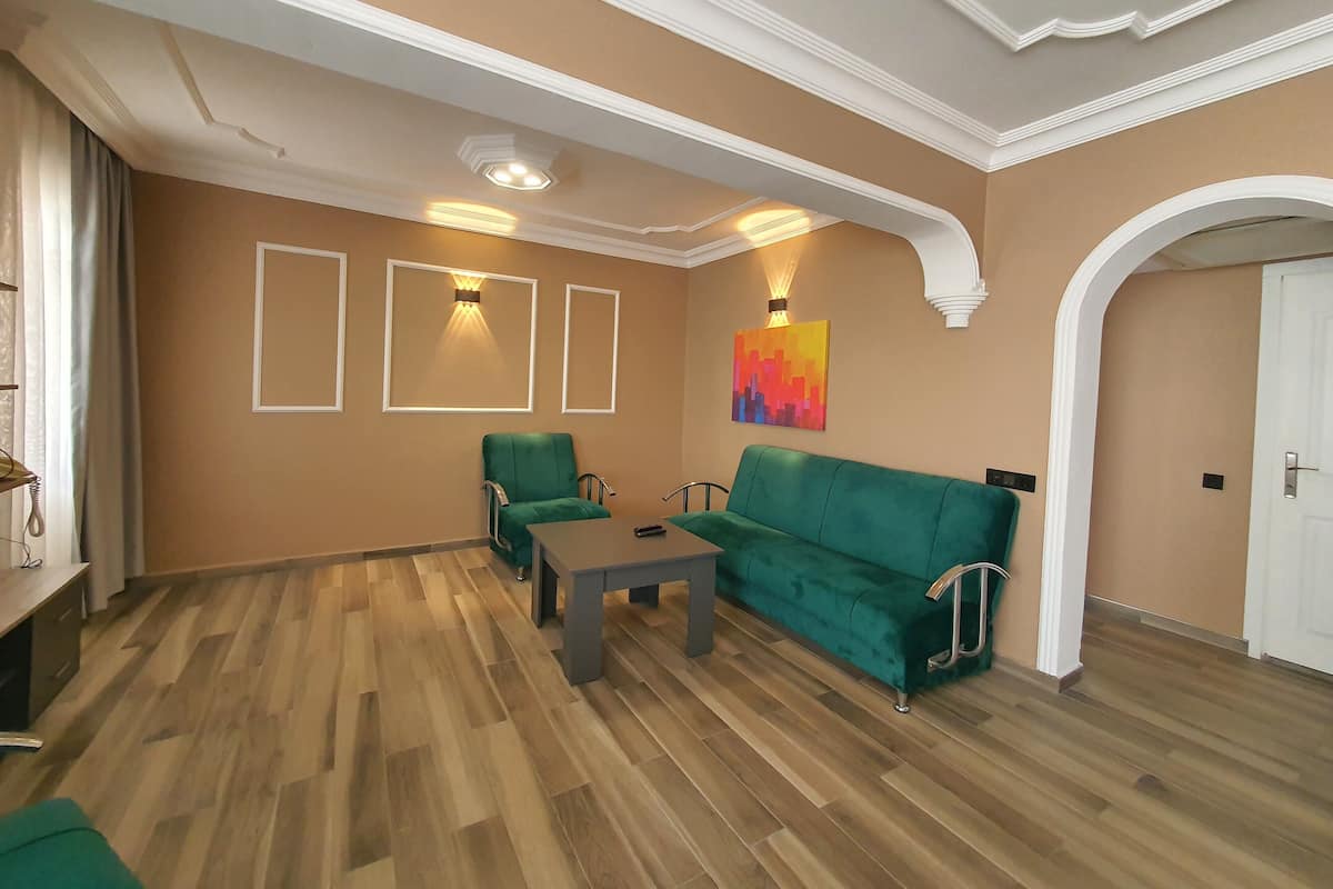 A living room with a green sofa, a small table, and a chair. There are three wall-mounted lights and a colorful painting on the wall.