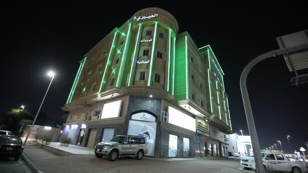 Un hôtel à plusieurs étages, doté d’éléments décoratifs verts illuminés, d’une entrée imposante et d’un espace de stationnement.