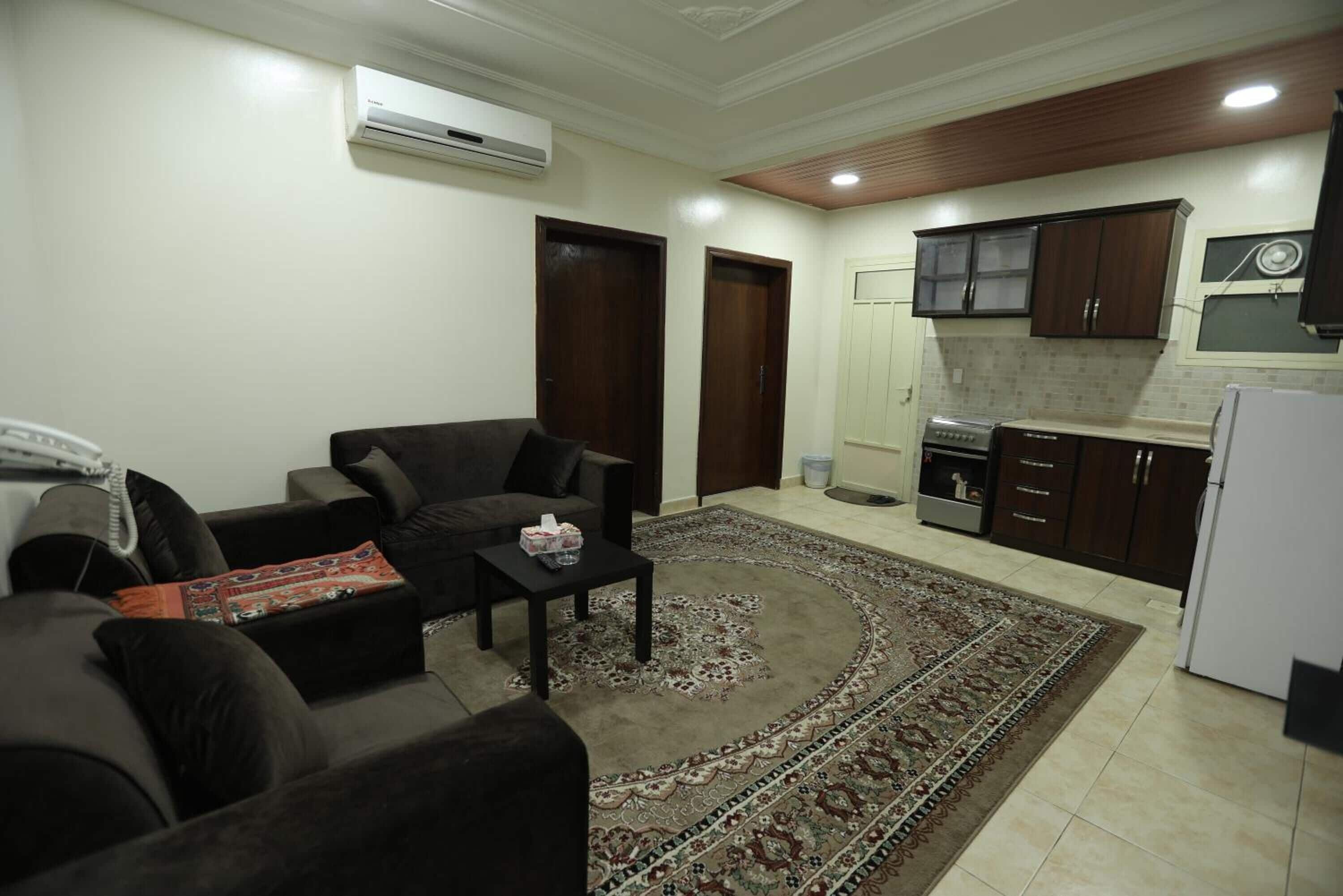 A living room with a sofa, a coffee table, and a patterned rug. There is a kitchen area with cabinets and a refrigerator.