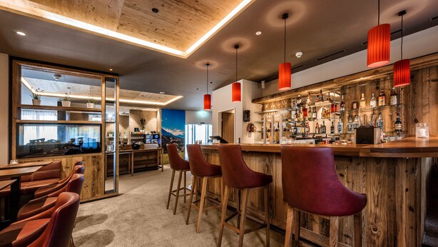 Een bar met een houten toog, rode beklede barkrukken en uitzicht op de keuken.