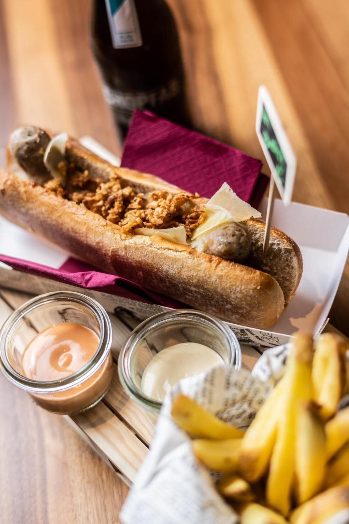 Ein Hot Dog mit Belag, eine Flasche und Soßen auf einem Holztisch.