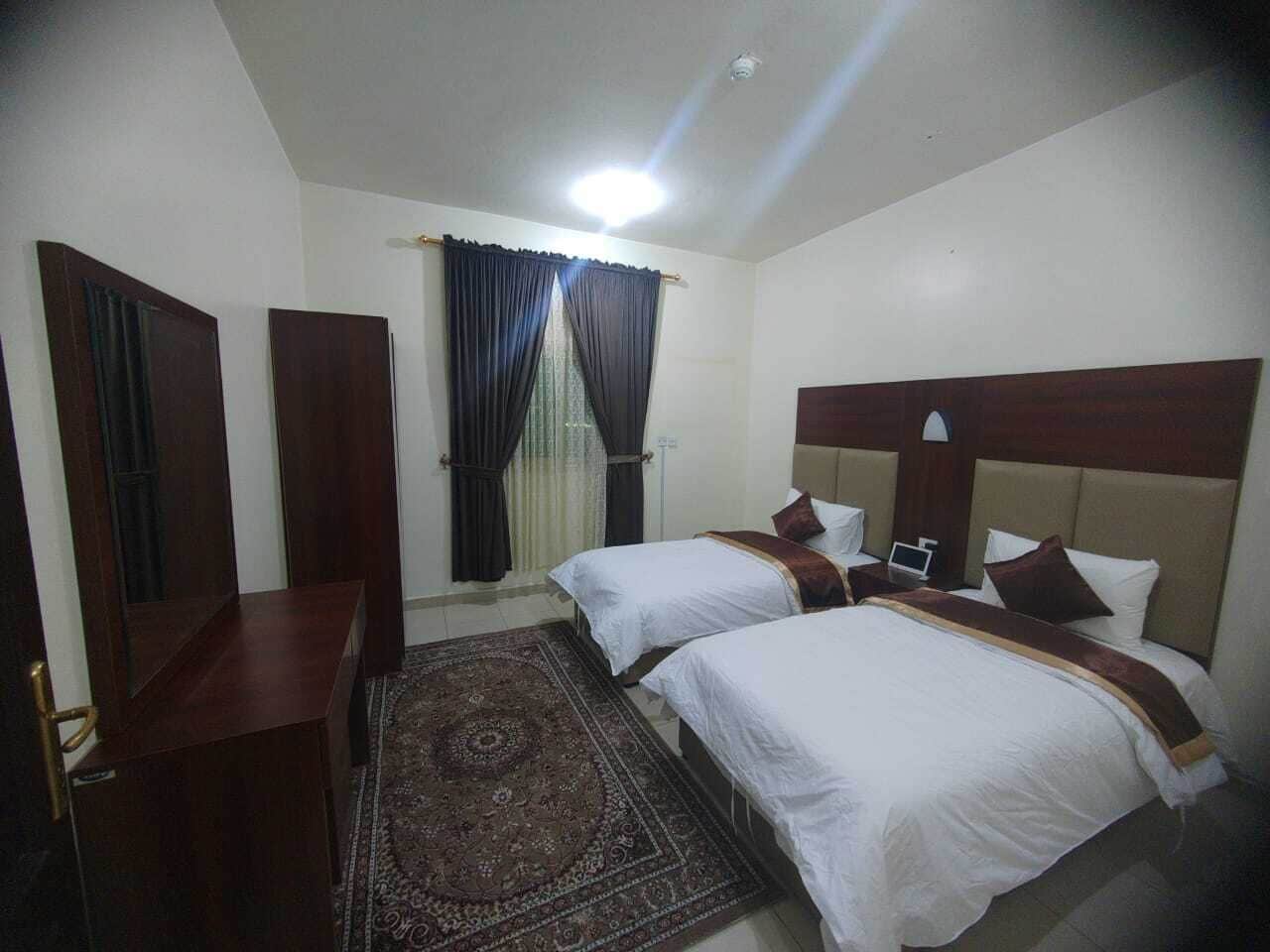 A hotel room with two beds, a desk, a chair, a window with curtains, and a patterned rug.