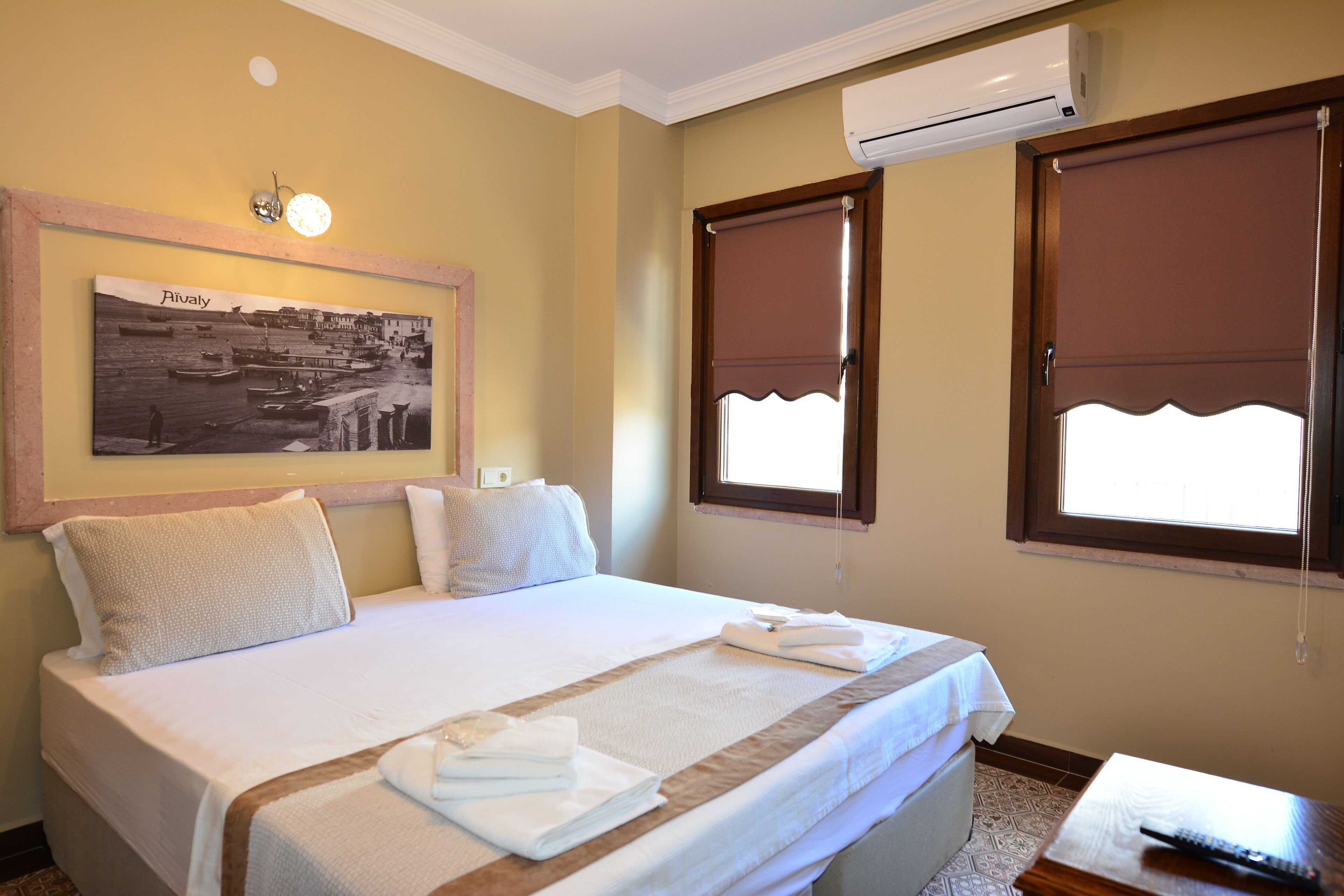 A neatly made bed with white linens and towels, a framed picture on the wall, and two windows with brown blinds.