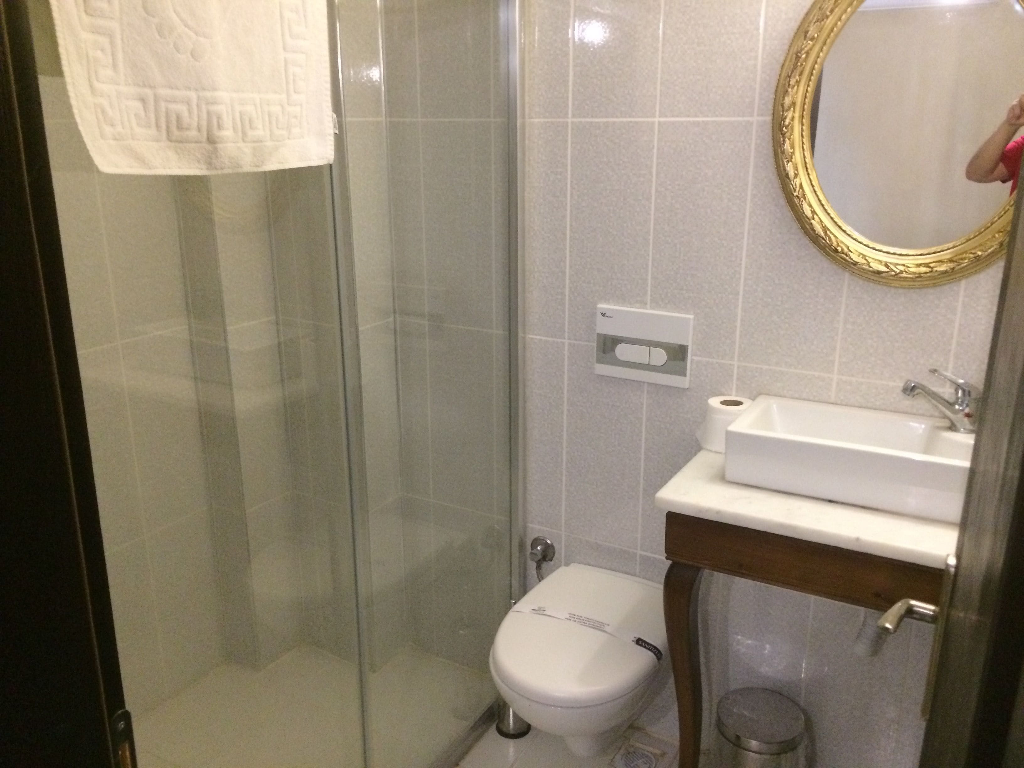 Un baño con cabina de ducha de vidrio, inodoro blanco, lavamanos con espejo y una toalla colgada en la pared.