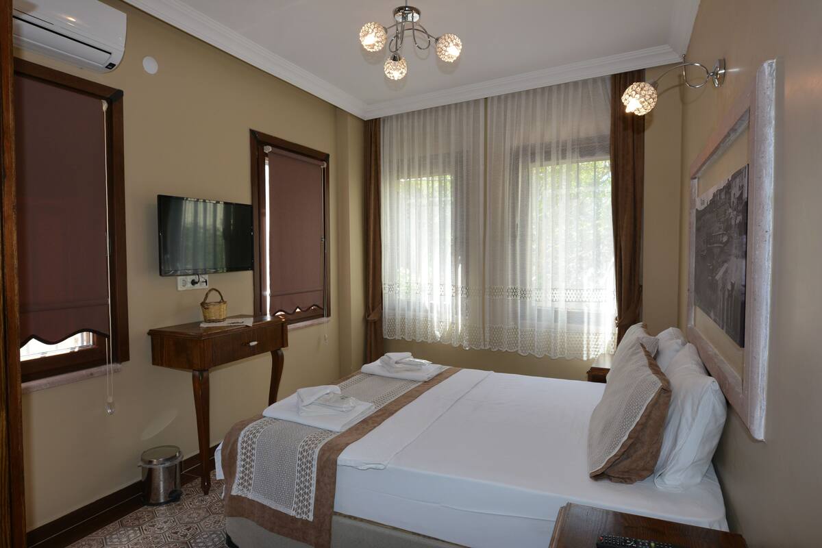 A hotel room with a bed, a television, a small table with a basket, a window with curtains, and an air conditioning unit.