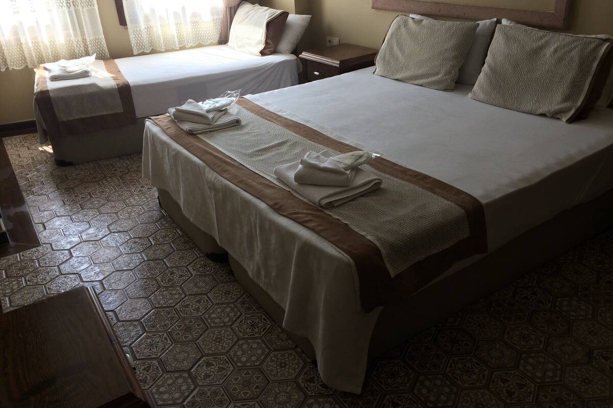A hotel room with two beds, each with neatly folded towels and a brown bed runner. The room has patterned hexagonal floor tiles and a framed picture on the wall.