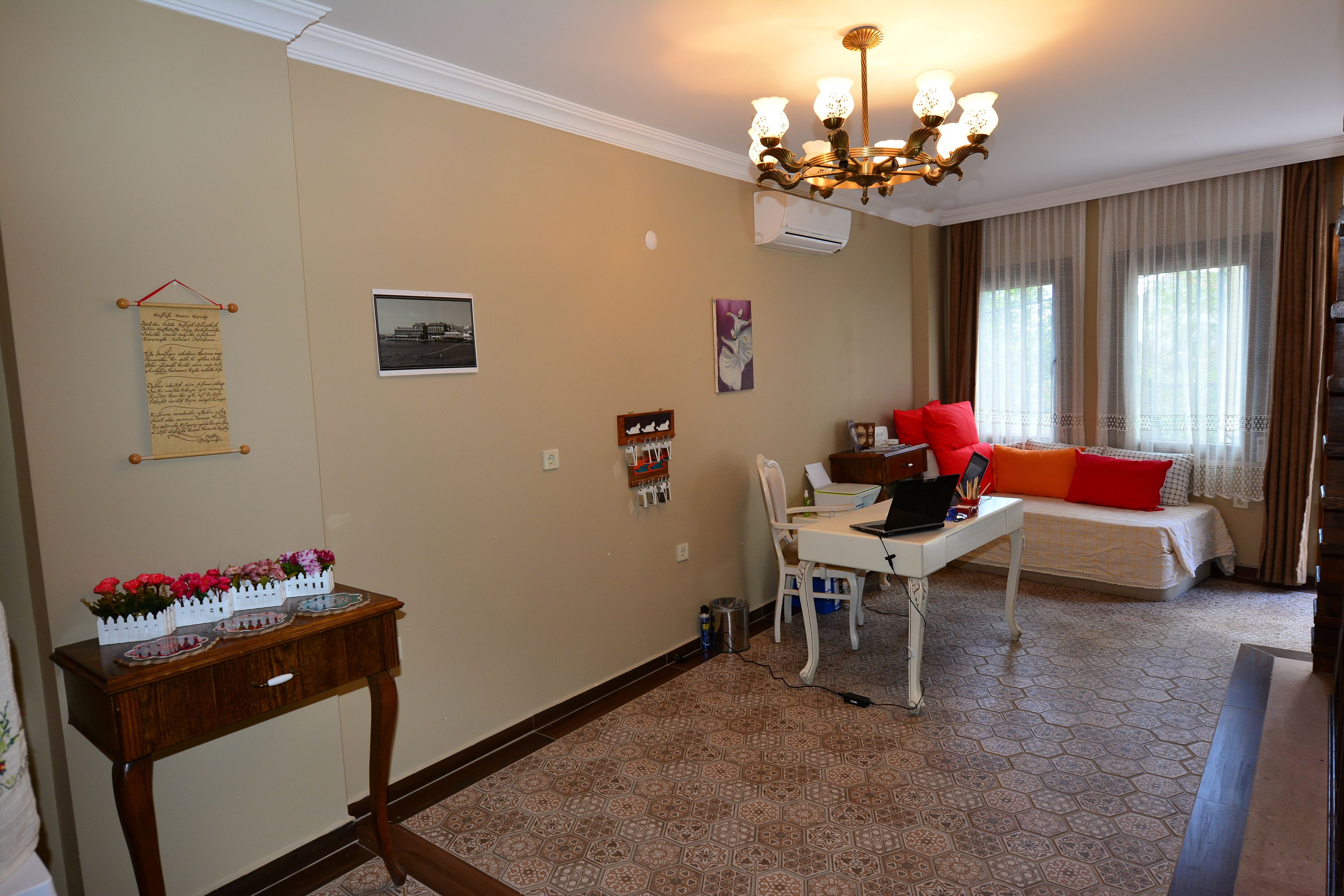 A room with a desk, chair, and a bed with red pillows. There is a chandelier, a wall-mounted air conditioner, and a window with curtains.