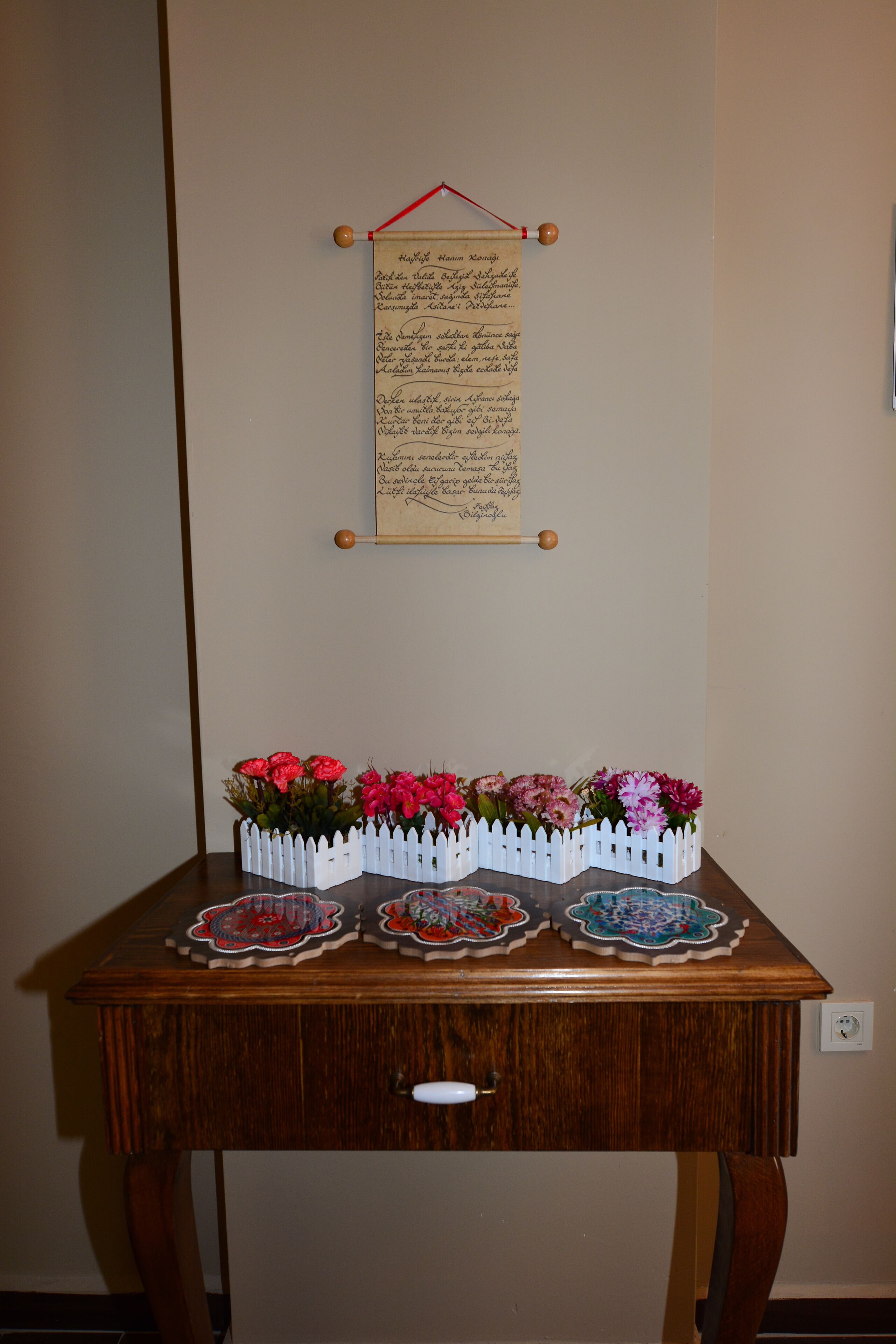 A wooden table with a white picket fence holding pink flowers, a handwritten note on a string, and a white switch on the wall.
