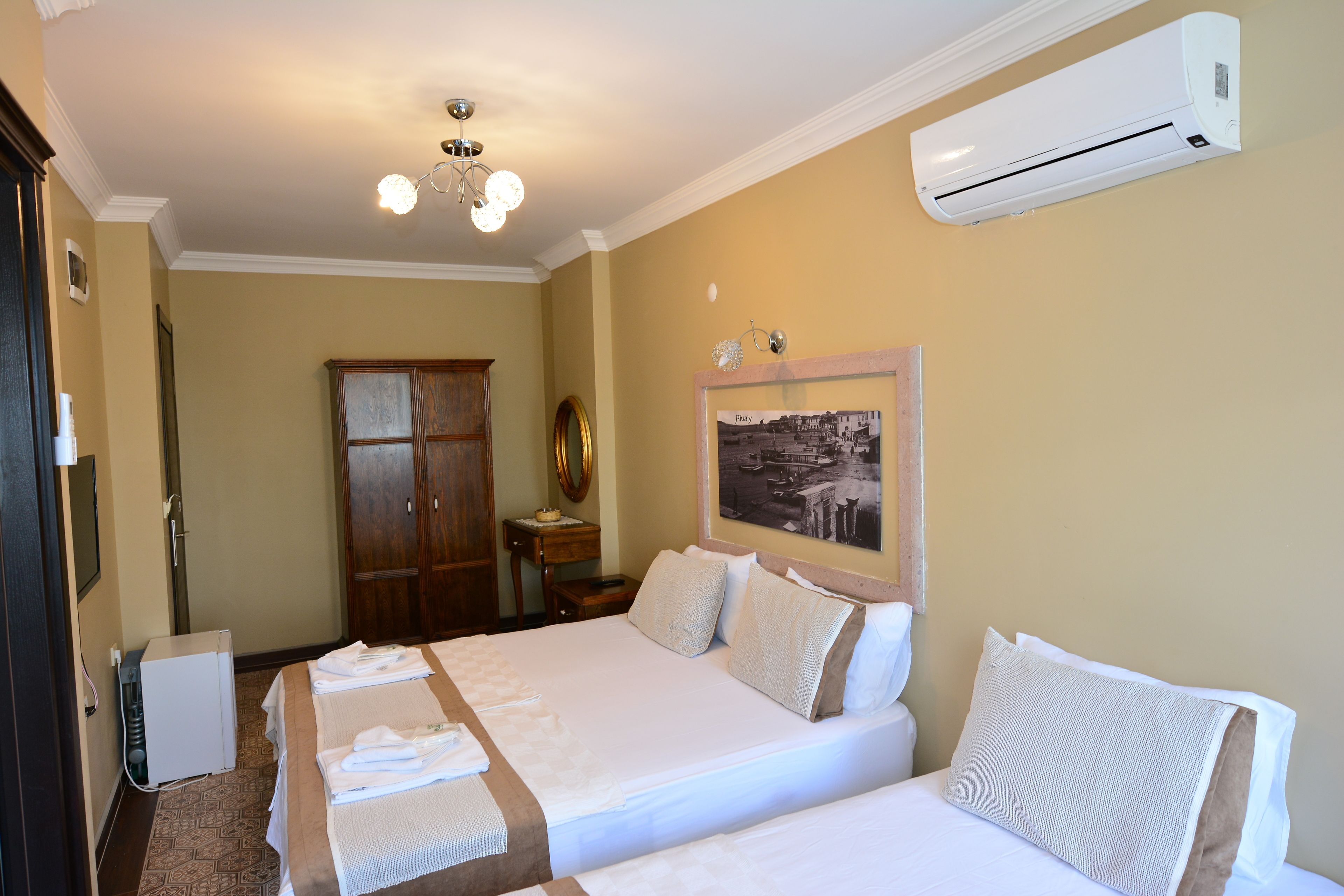 A hotel room with two beds, a ceiling fan, an air conditioner, a wooden wardrobe, a small table, and a framed picture on the wall.
