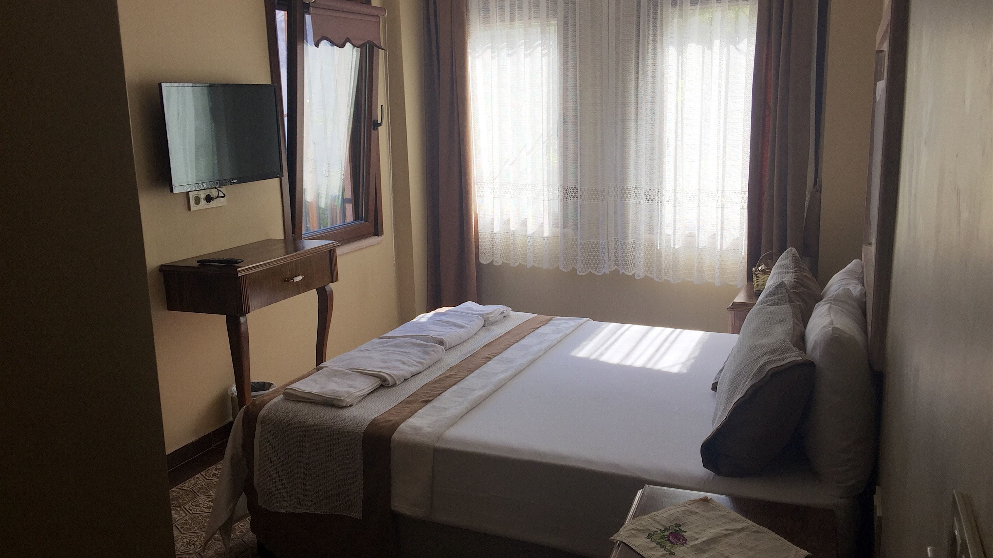 Una camera d'albergo con un letto, una TV a parete, una finestra con tende e un tavolino con un cesto.