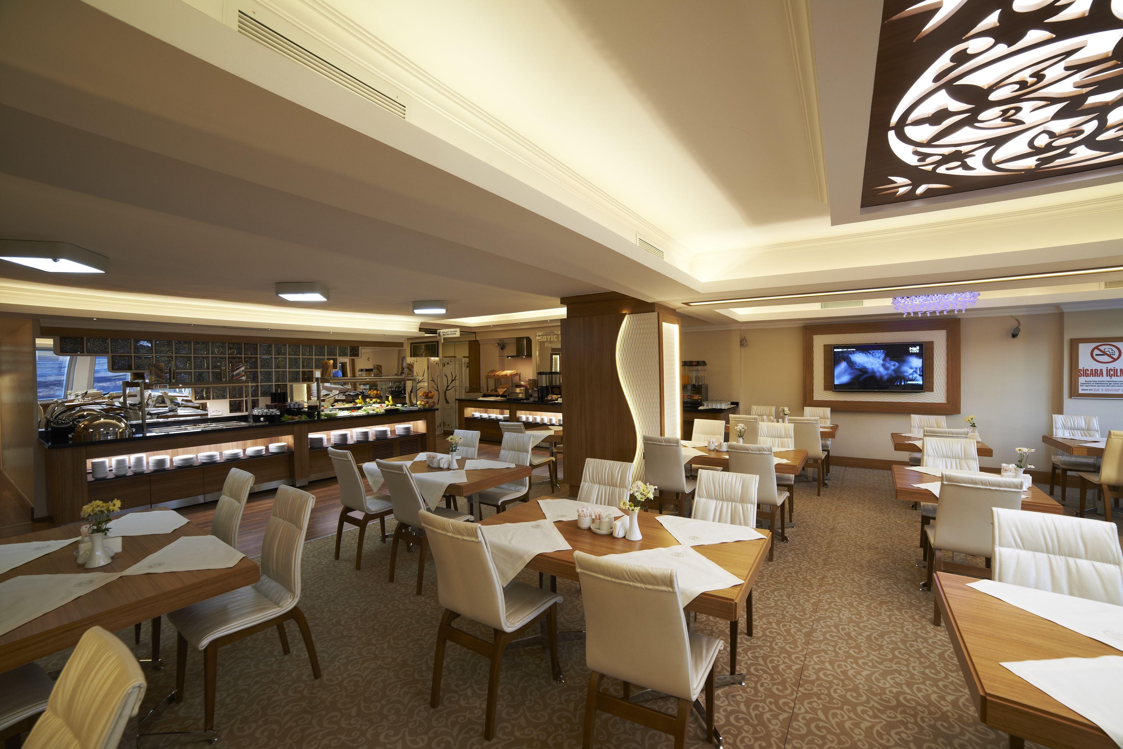 A hotel dining area with multiple tables and chairs, a buffet counter, and a television screen.