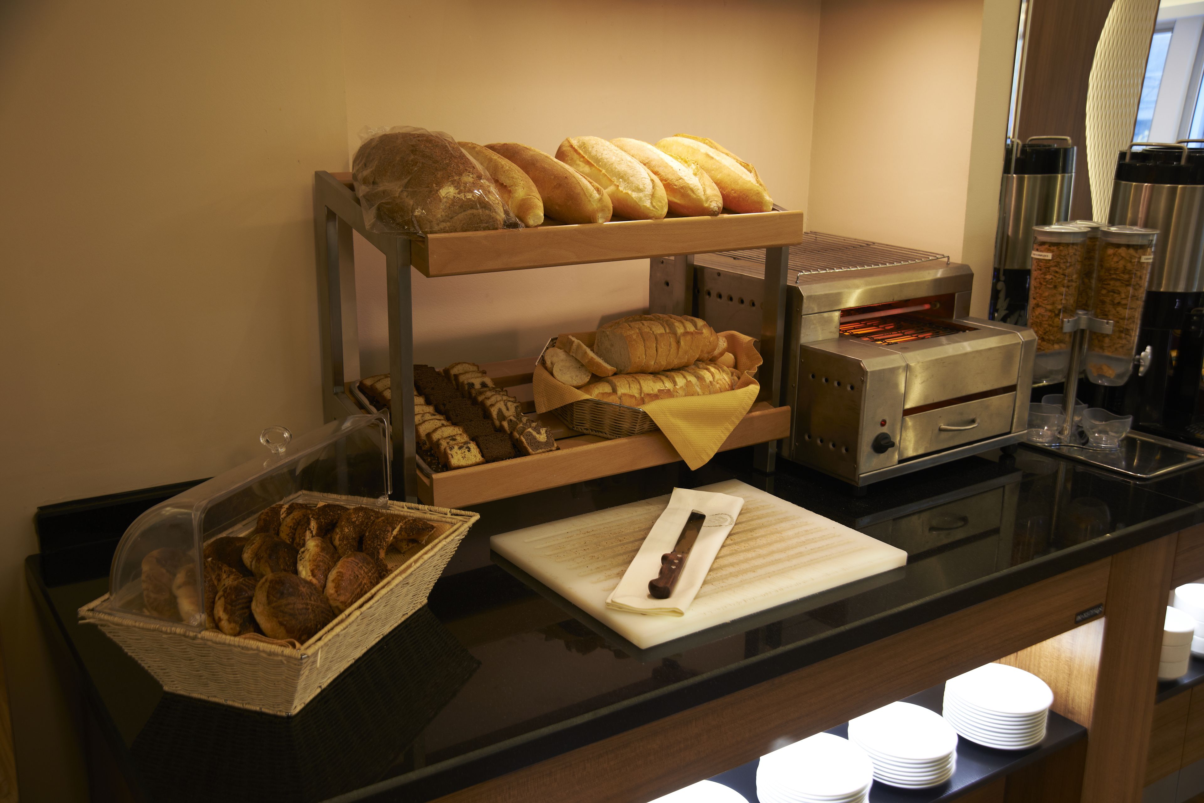 A buffet counter with various breads and pastries displayed on shelves, a bread slicer, and a coffee machine.
