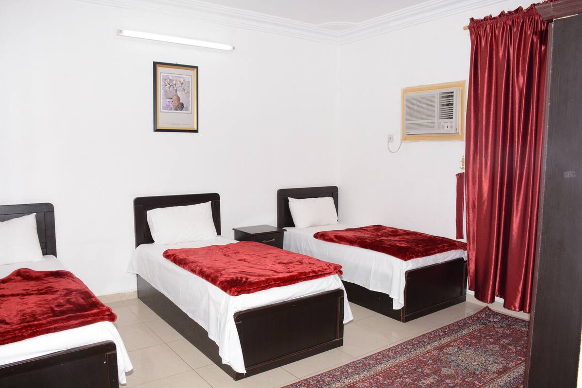 Une chambre avec deux lits, chacun recouvert d’une couverture rouge et de draps blancs, un petit tableau encadré accroché au mur et un rideau rouge à la fenêtre.
