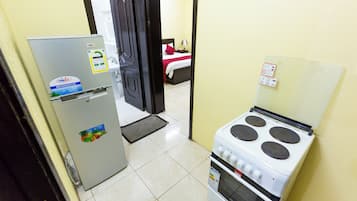 Cozinha compacta com geladeira, fogão e uma cama visível por uma porta aberta.