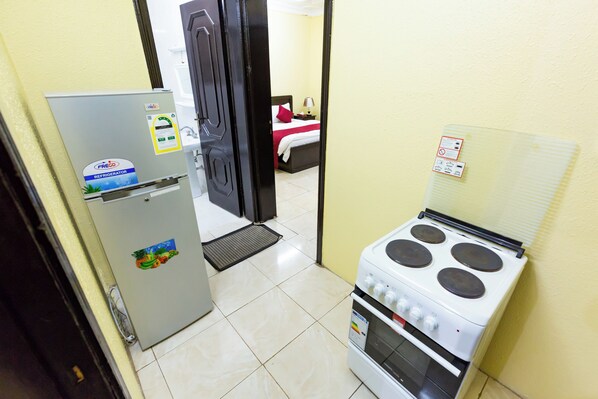 A compact kitchen with a refrigerator, stove, and a bed visible through an open door.