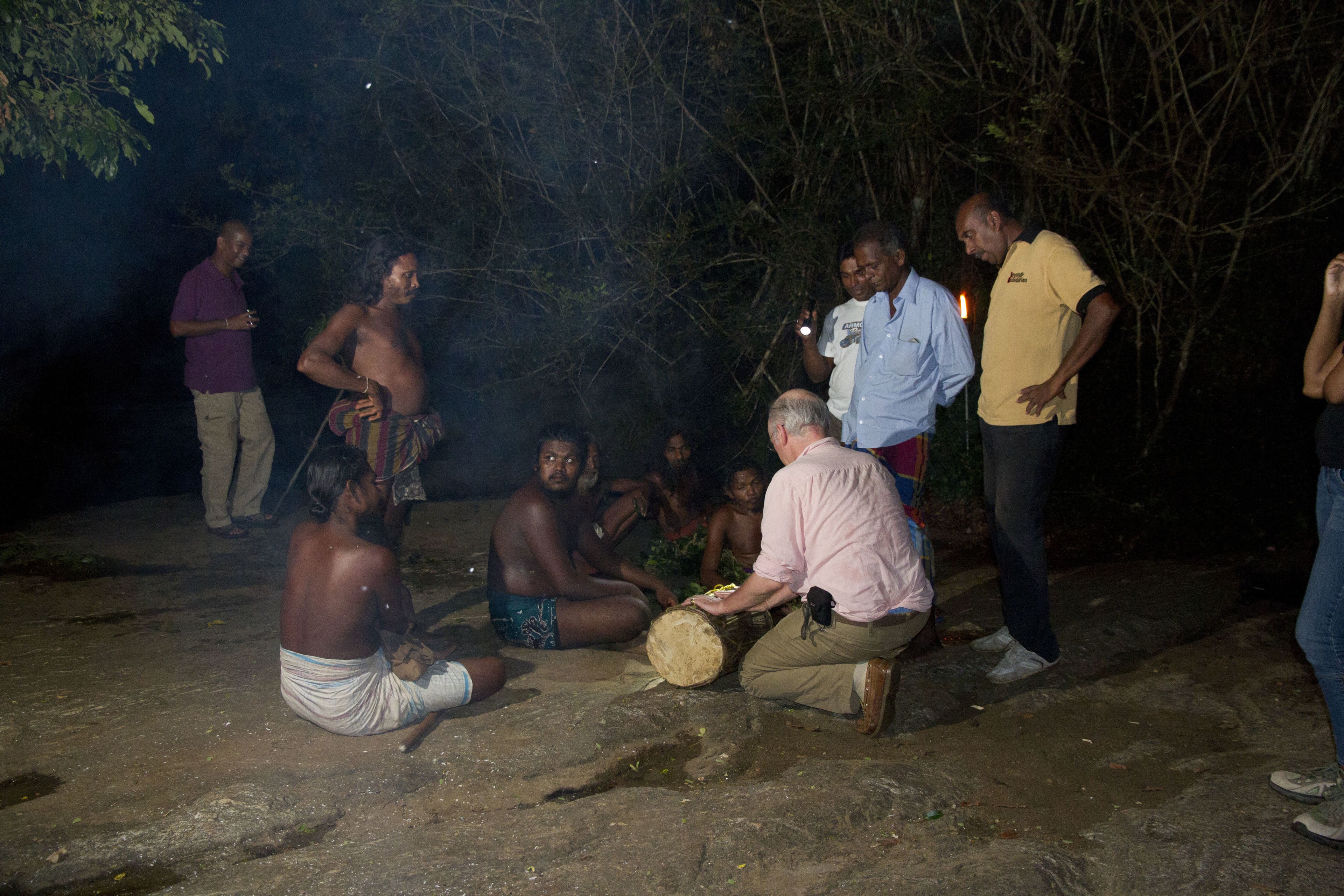 A group of people gathered around a drum in a natural setting at night.