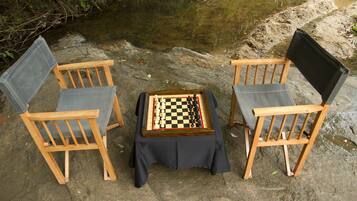 Two wooden chairs with grey cushions and a chessboard on a table, set on a stone surface near a stream.
