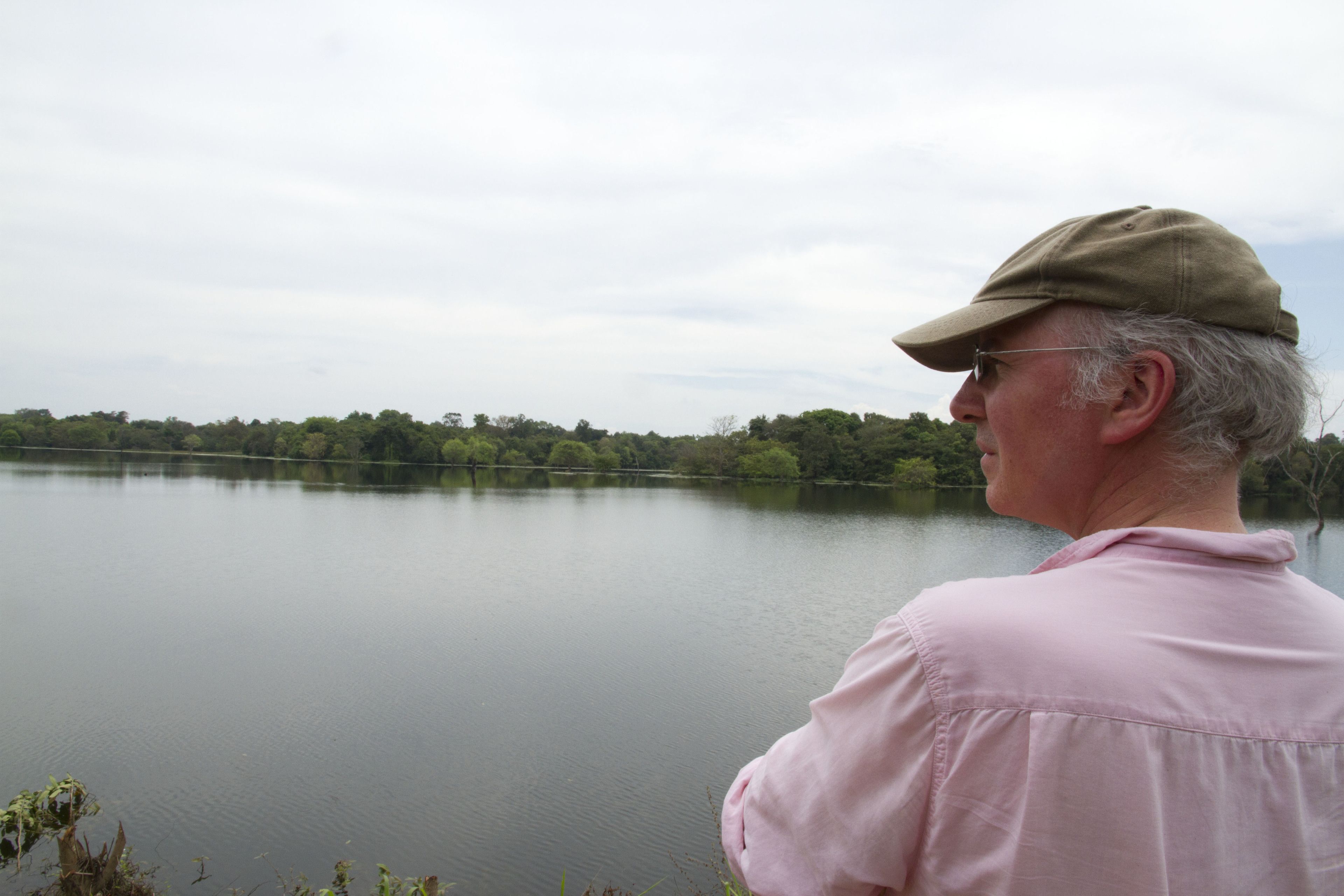 Una persona de pie junto a un lago tranquilo con árboles al fondo.