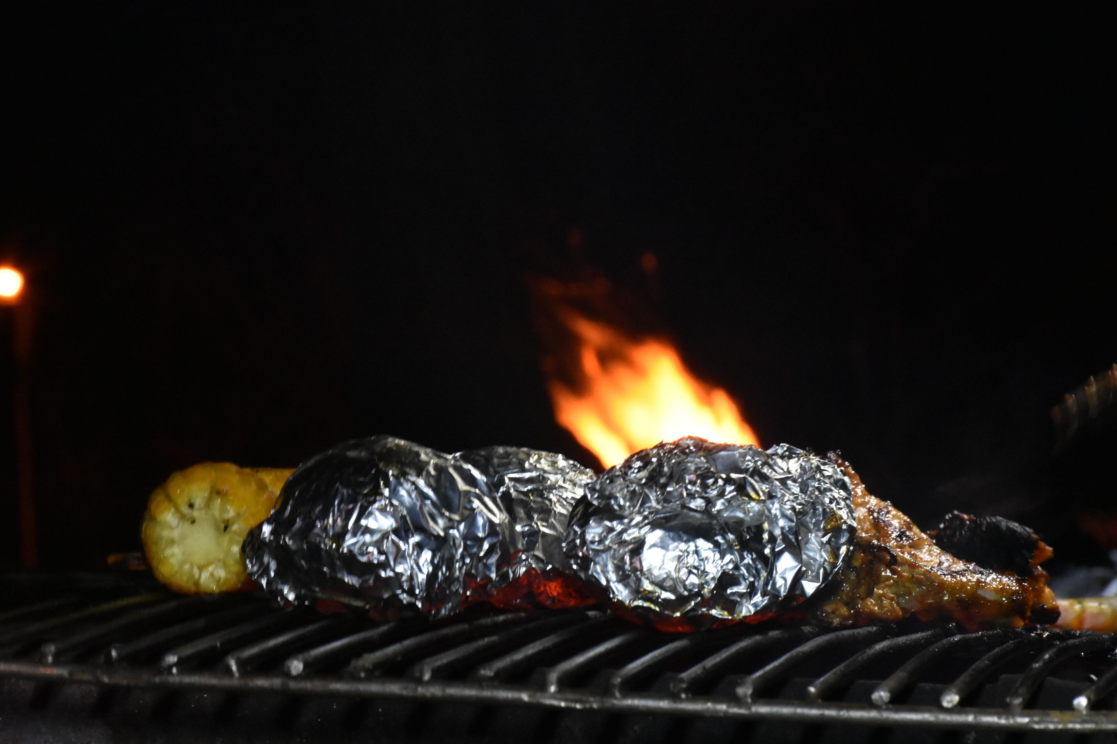 Una parrilla con alimentos, incluyendo un trozo de carne y una patata, con fuego al fondo.
