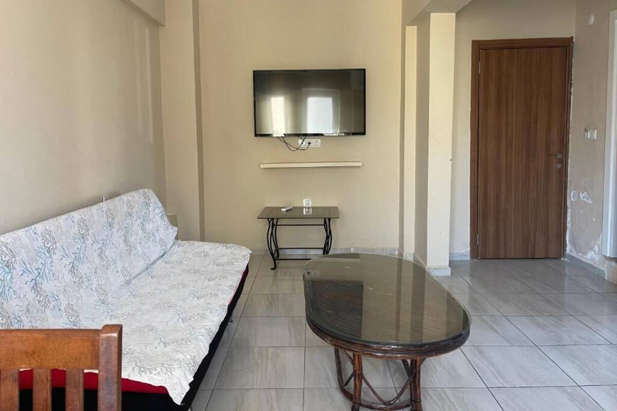Una sala de estar con un sofá, una mesa con cubierta de vidrio, un televisor de pantalla plana montado en la pared y una puerta de madera al fondo.