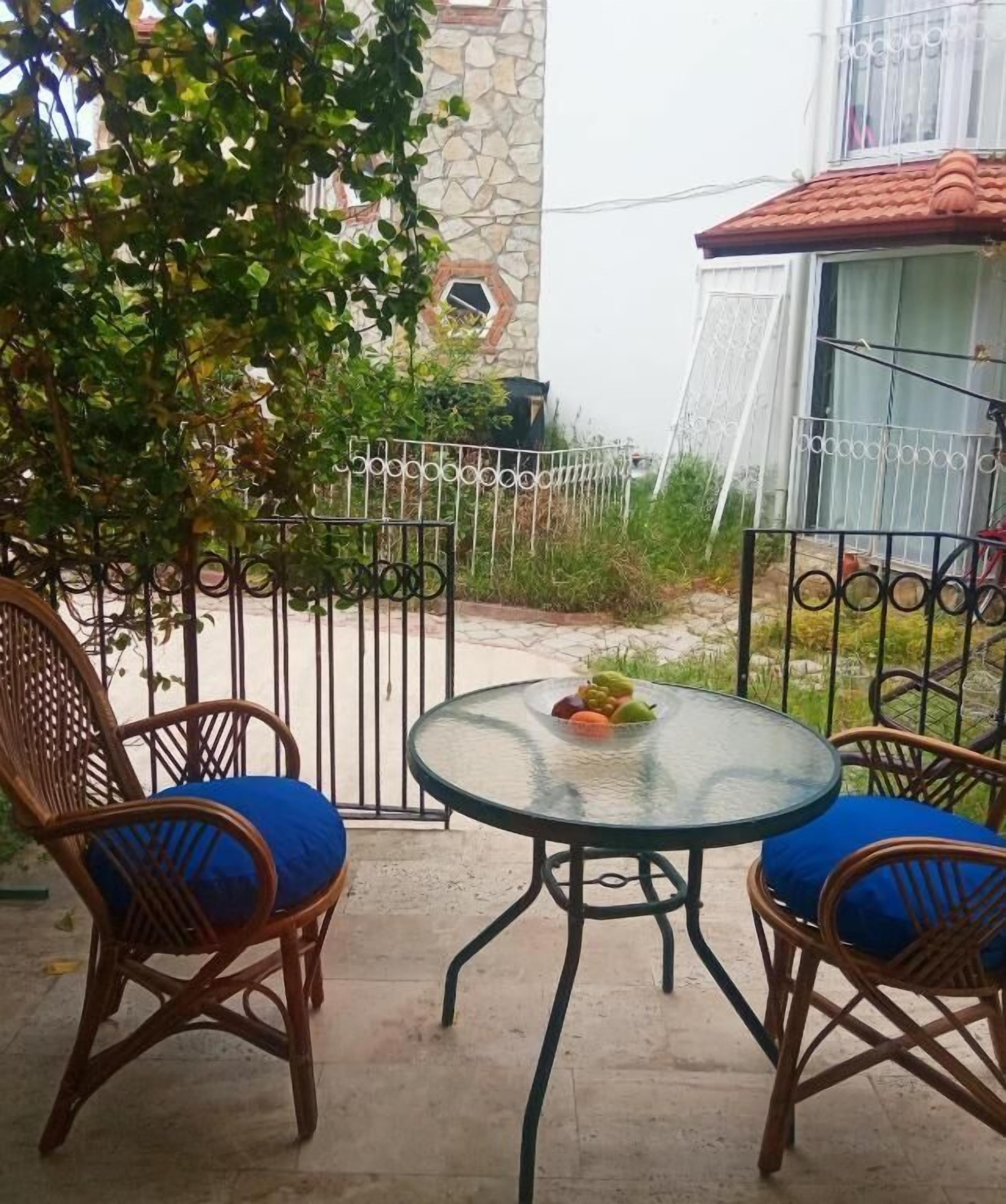 A patio with two wicker chairs and a glass table, surrounded by a metal fence and a stone wall.