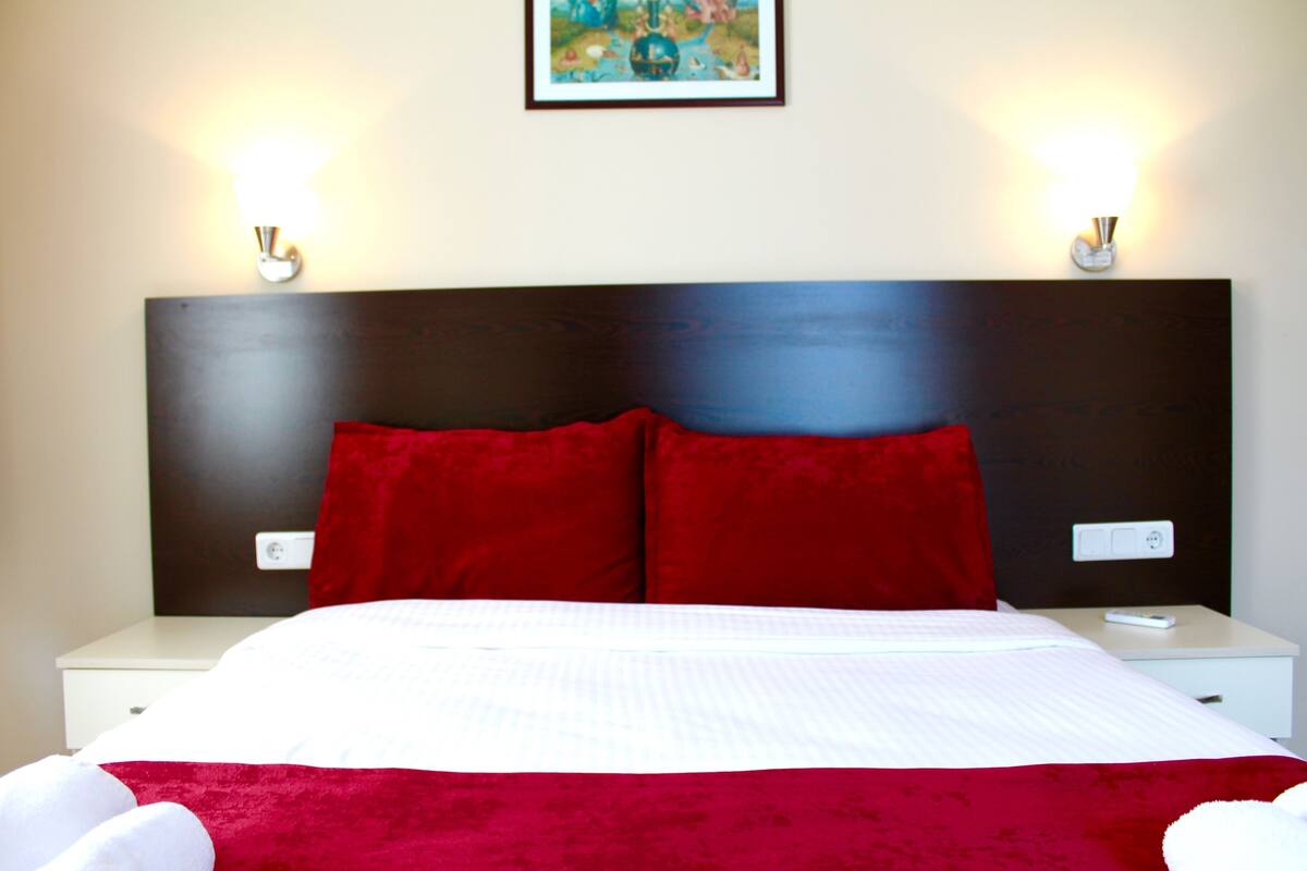 Una cama bien hecha con una colcha blanca y roja, dos almohadas rojas y un cabecero oscuro.