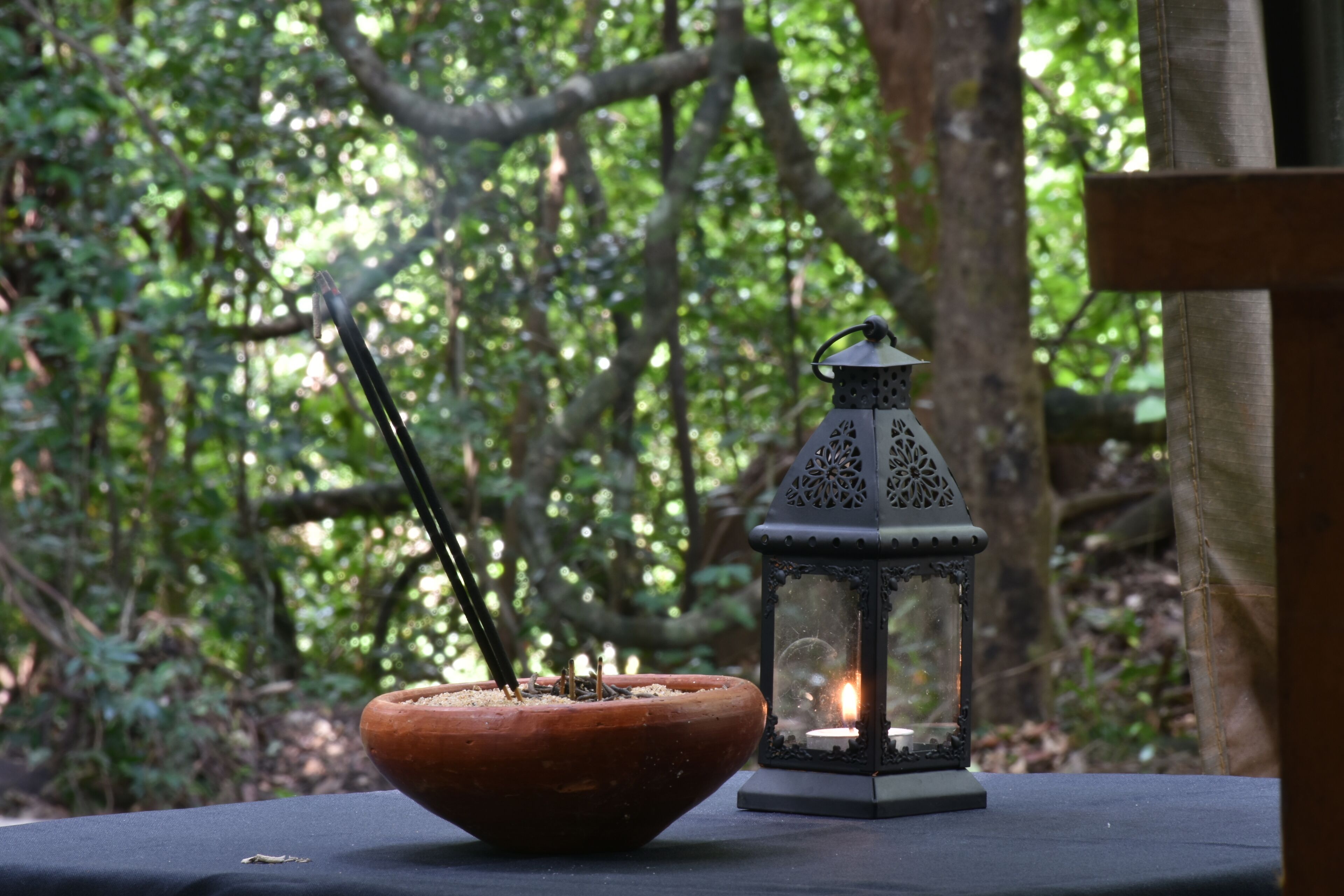 A candle in a bowl on a table with a lantern in a forest setting.