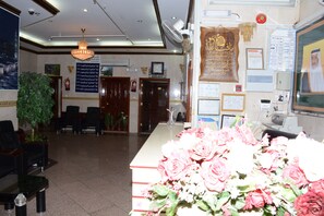 Uma área de recepção com balcão, cadeiras, um candelabro, uma planta e diversos cartazes informativos.
