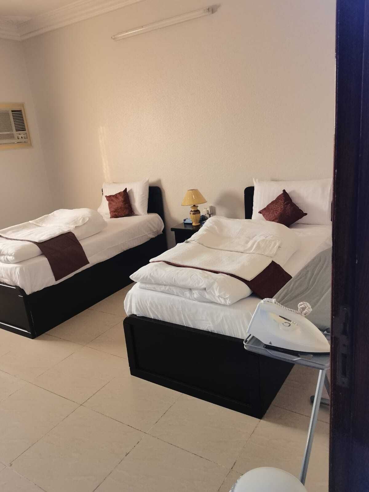 Habitación de hotel con dos camas, cada una con ropa blanca y almohadas marrones. Hay una mesita de noche con una lámpara entre las camas.