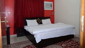 Een kamer met een bed, nachtkastje, rood gordijn, een wandairconditioner en een kamerbreed tapijt met dessin.