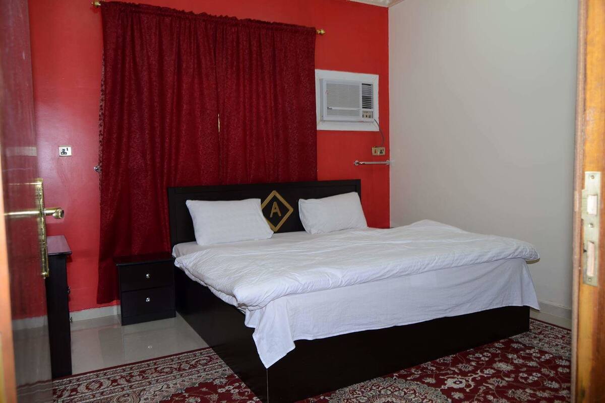 Une pièce comprenant un lit, une table de chevet, un rideau rouge, un climatiseur fixé au mur et un tapis à motifs.