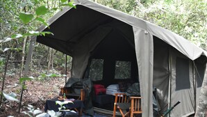 Una tienda de campaña montada en un bosque, con una mesa, sillas y una maleta roja.