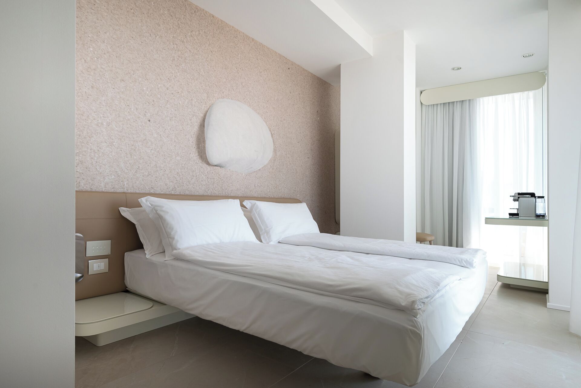 A modern hotel room with a large bed, a built-in bench, a large window with curtains, and a wall-mounted light fixture.