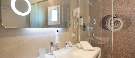 A modern bathroom with a white sink, a mirror with a circular light, a handheld hairdryer, and a glass shower enclosure.