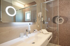 A modern bathroom with a white sink, a mirror with a circular light, a handheld hairdryer, and a glass shower enclosure.