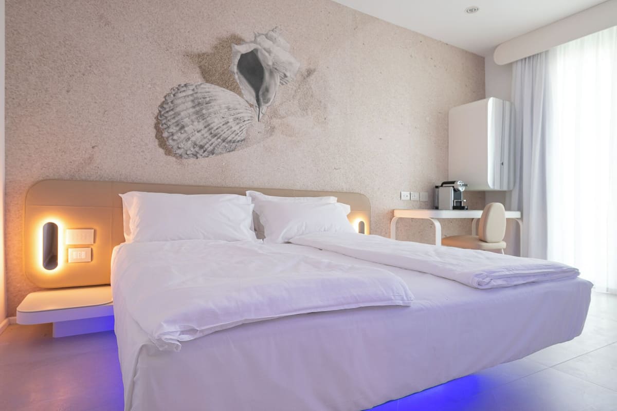 A modern hotel room with a large bed, a wall-mounted light, a small desk, and a chair.