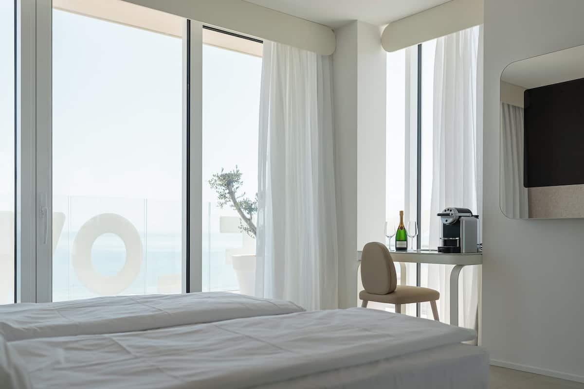 A hotel room with a large window, a bed, a desk with a chair, and a minibar.