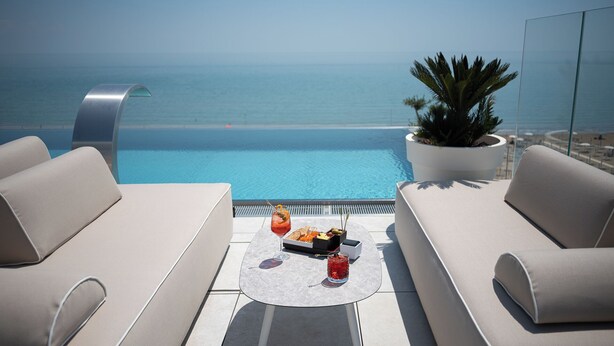 A rooftop terrace with a pool, two sofas, a table with drinks, and a potted plant.