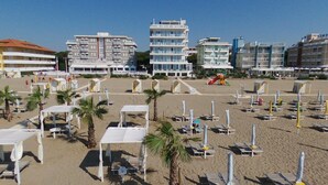 A beach with rows of sun loungers and umbrellas, a children's play area, and several multi-story buildings in the background.